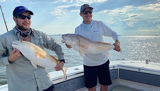 Two men stand on a boat with the large redfish they caught on a Galveston jetty fishing trip.